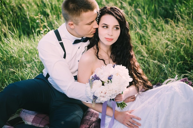 Recém-casados apaixonados estão sentados na grama no parque a noiva e o noivo sorriem e se divertem no piquenique do casamento