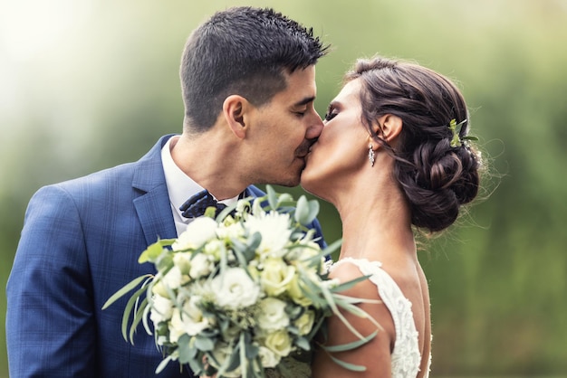 Recém-casados beijam ao ar livre enquanto seguram um buquê de flores brancas na frente deles