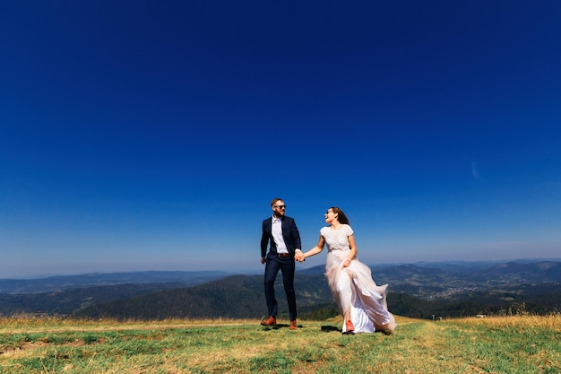 Recém-casados dão as mãos e correm na grama verde no topo da montanha