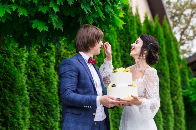 Recém-casados e damas de honra se divertem e comem bolo de casamento juntos ao ar livre em um banquete de casamento.