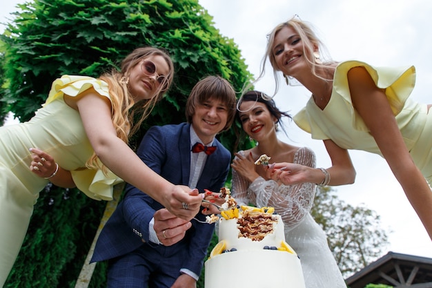 Recém-casados e damas de honra se divertem e comem bolo de casamento juntos ao ar livre em um banquete de casamento.