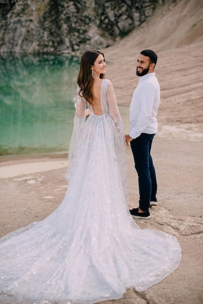 Foto recém-casados estão de pé perto de um belo lago. noivo do oriente médio e noiva caucasiana de mãos dadas na praia. vista traseira.