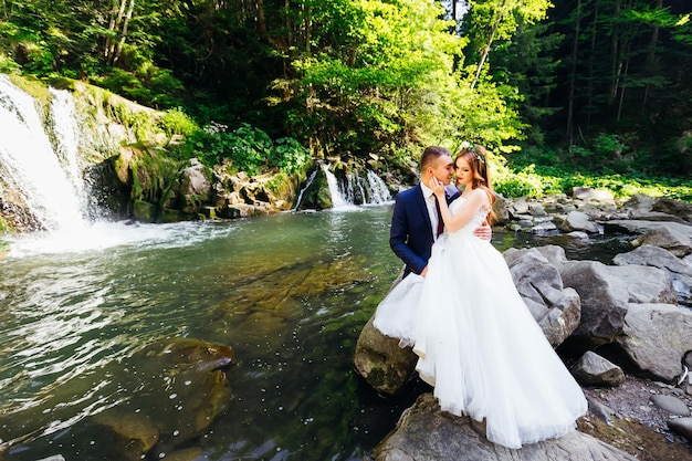 Recém-casados se abraçam ao fundo do rio e das montanhas enquanto estão sentados em uma grande pedra
