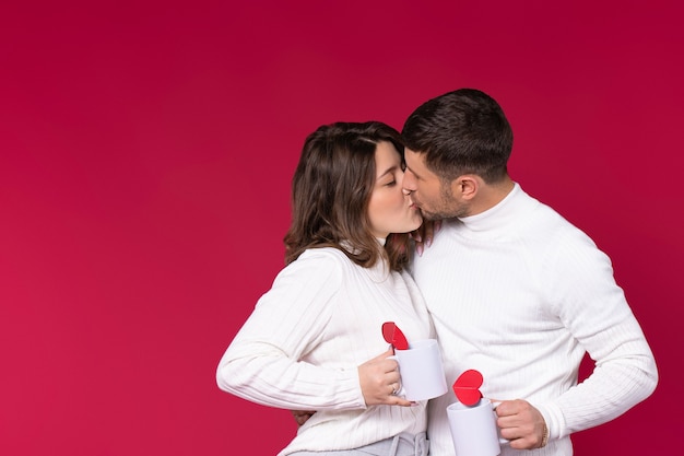 Recém-casados se beijando em um fundo vermelho segurando copos brancos com corações feitos à mão.