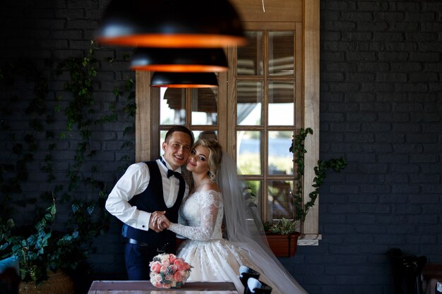 Recém-casados sorridentes abraçando perto da janela interior. retrato de noivos no quarto de hotel elegante. casal de noivos em um quarto com um interior elegante com lâmpadas. dia do casamento