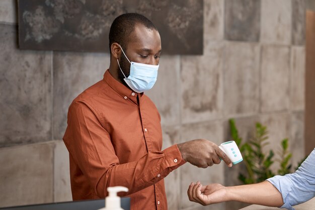 Recepcionista africano usando máscara médica e termômetro infravermelho para medir o corpo