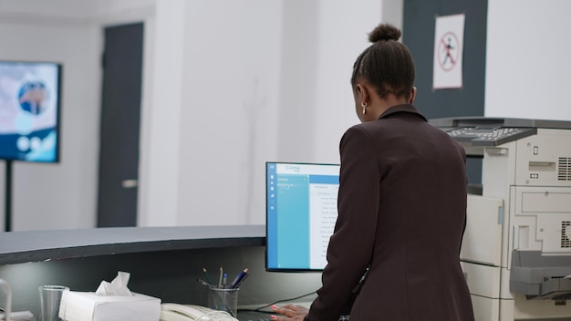 Recepcionista afroamericana que trabaja en una computadora con citas e informes médicos en el mostrador de registro del hospital. Trabajador de la clínica mirando las visitas de control en el monitor en el mostrador de recepción.