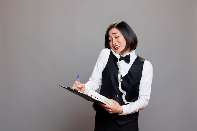 Una recepcionista asiática sonriente hablando con un cliente en un teléfono fijo y escribiendo en el clipboard. Una joven camarera atractiva en uniforme teniendo una conversación amistosa en un teléfono retro