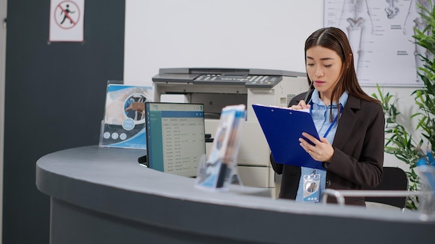 Recepcionista que trabaja en citas de atención médica con informes de revisión en el mostrador de recepción del hospital. Uso de formularios de consulta para brindar soporte de seguros y ayuda con los exámenes en el mostrador.