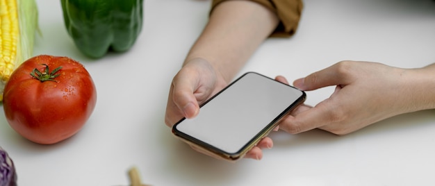 Receta de aspecto femenino en el teléfono inteligente de la pantalla en blanco mientras se prepara el ingrediente para cocinar en la mesa de la cocina con verduras frescas