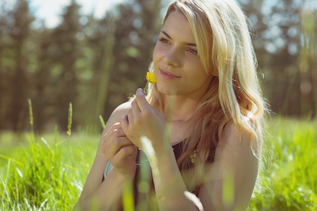 Recht blond in den sundress, die auf dem Gras sitzt, das gelbe Blume hält