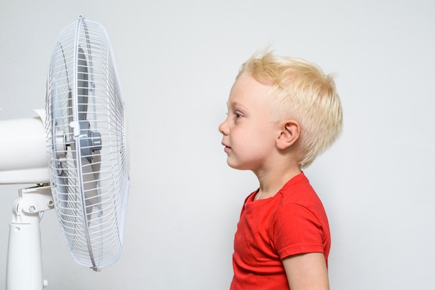 Recht blonder Junge im roten Hemd steht nahe einem Ventilator.