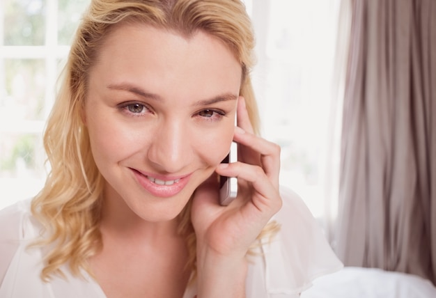 Recht blondes Sitzen auf Bett am Telefon, das an der Kamera lächelt