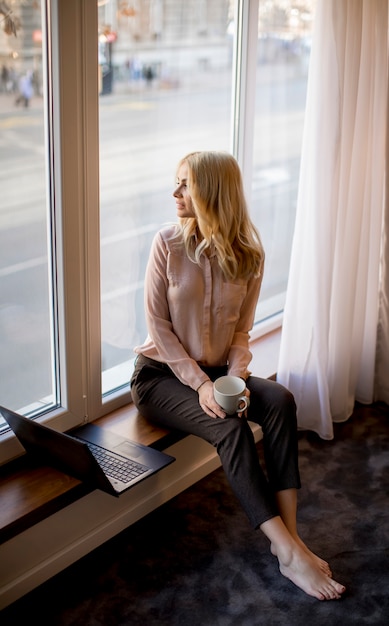 Recht junge Frau, die Laptop verwendet und durch Fenster sitzt