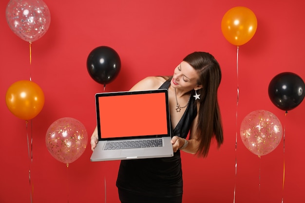 Recht junge Frau im schwarzen Kleid, das auf Laptop-PC-Computer mit leerem schwarzem leerem Schirm in den Händen auf hellroten Hintergrundluftballonen schaut Spott oben. Frohes neues Jahr, Geburtstagsmodell Urlaub Partykonzept.