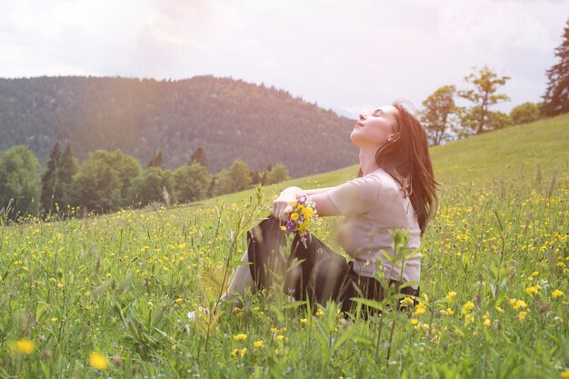 Recht junge Frau mit Blumenstrauß von den Wildblumen, die den Rasen sitzen. Berge auf der. Sommertag sonnig