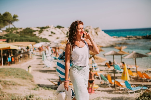 Recht junge Frau mit einer Tasche unter Verwendung des Handys auf dem Strand