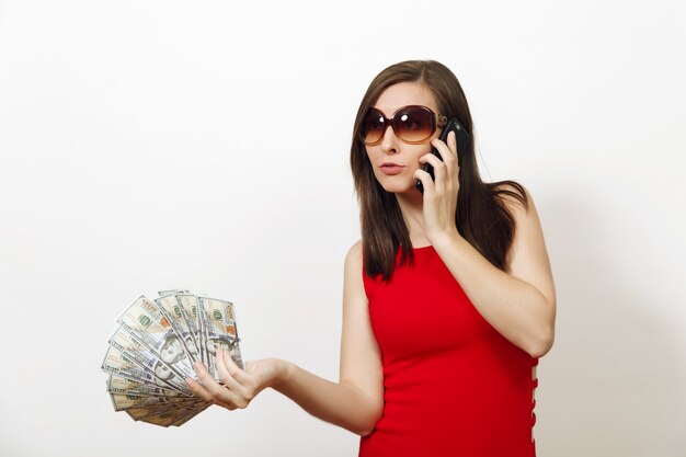 Recht kaukasische junge glückliche Geschäftsfrau im roten passenden Kleid und in den Gläsern, die auf dem Handy sprechen, Geldbanknoten auf weißem Hintergrund halten. Schönes Mädchen mit Bargeld, das beiseite lokalisiert schaut.