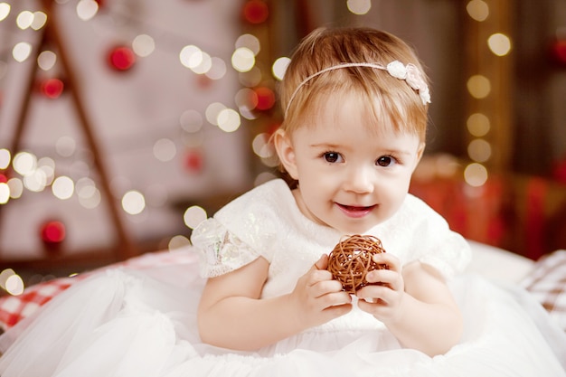 Recht kleines Mädchen im weißen Kleid, das über Weihnachtsbaum und Lichter spielt und glücklich ist.