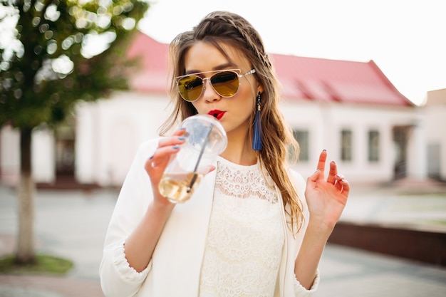 Recht modisches Mädchen mit Borte in widergespiegelter Sonnenbrille Kaffee in der Straße trinkend.