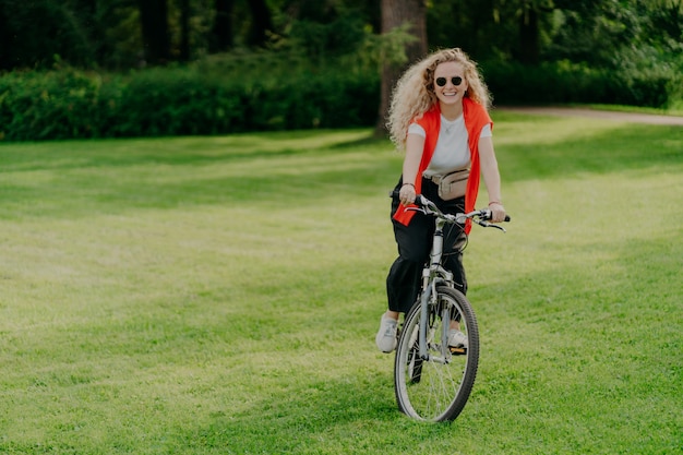 Recht nette junge Frau reitet Fahrrad, trägt Sonnenbrille, Freizeitkleidung, wirft auf grünem Rasen auf