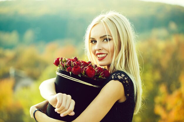 Recht nettes Mädchen oder schöne blonde Frau im sexy schwarzen Kleid mit roten Rosenblumen im Kasten im Freien o