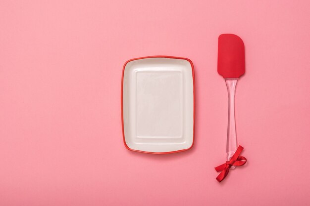 Rechteckiger Teller und Küchenspatel auf rosa Hintergrund. Küchengeräte auf festlichem Hintergrund. Flach liegen.