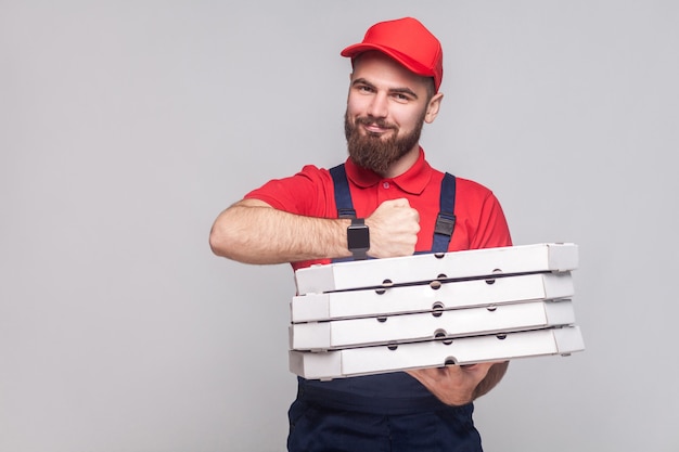 Rechtzeitig! Junger fröhlicher Lieferbote mit Bart in blauer Uniform und rotem T-Shirt stehend, mit Stapel Pizzakartons aus Pappe auf grauem Hintergrund. Innen, Studioaufnahme, isoliert, Kopienraum.