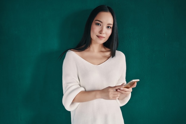 Recibiendo buenos mensajes. Hermosa mujer joven con teléfono inteligente y sonriendo mientras está de pie contra la pared verde