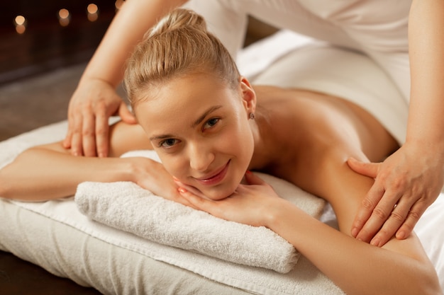 Recibir masaje relajante. Sonriente a guapa chica desnuda acostada sobre toallas blancas mientras el maestro de masajes procesa sus hombros