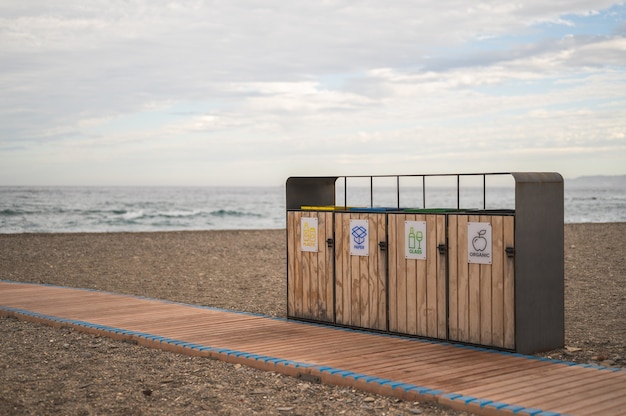 Reciclaje de contenedores frente a un camino de madera en la playa con el mar detrás y el cielo nublado. concepto de conservación del medio ambiente