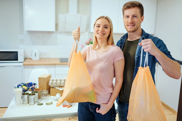 Reciclaje. Responsable joven sonriente familia sosteniendo bolsas de basura mientras está de pie cerca de la mesa llena de desechos en casa