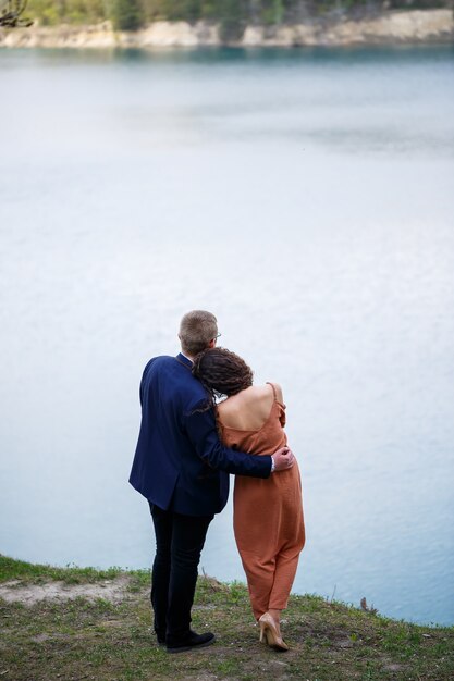 Recién casados alegres van tomados de la mano y riendo, en el contexto de un lago y un prado verde. Novio alegre y hermosa novia con pelo rizado a pie en la pradera