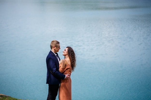 Recién casados alegres van tomados de la mano y riendo, en el contexto de un lago y un prado verde. Novio alegre y hermosa novia con pelo rizado a pie en la pradera