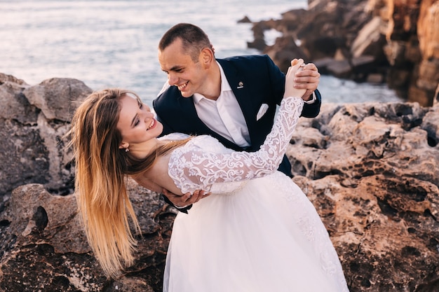 Recién casados bailan en la costa rocosa con vista al mar