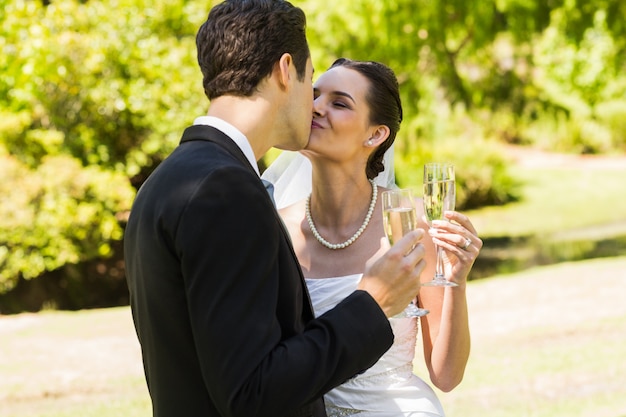 Recién casados ​​besándose mientras tuestan flautas de champán en el parque