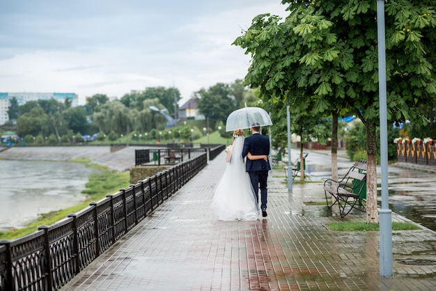 Los recién casados caminan bajo un paraguas bajo la lluvia en el parque