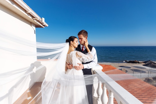 Los recién casados se disfrutan tiernamente a la sombra del velo volador en el fondo del hermoso mar