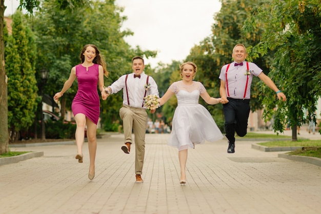 Los recién casados se divierten en el día de la boda junto con sus amigos, tomados de la mano que corren por la calle.