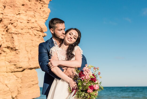 Los recién casados felices están parados tomados de la mano en el fondo del mar azul. Paseo nupcial en una playa de arena. En el fondo, cielo azul