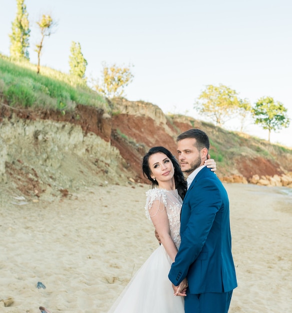 Los recién casados felices están parados tomados de la mano en el fondo del mar azul. Paseo nupcial en una playa de arena. En el fondo, cielo azul