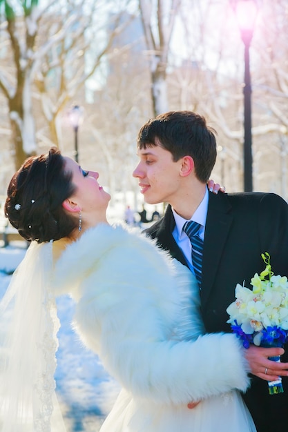 Recién casados de la joven pareja caminando en un bosque de invierno en la nieve. Novios abrazándose en el parque en invierno. Un hombre y una mujer hermosos con sus trajes de boda se encuentran entre los pinos.