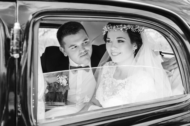 Recién casados con ropa festiva se sientan en un coche en fotografía en blanco y negro