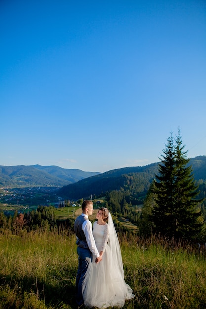 Los recién casados sonríen y se abrazan entre el prado en la cima de la montaña.