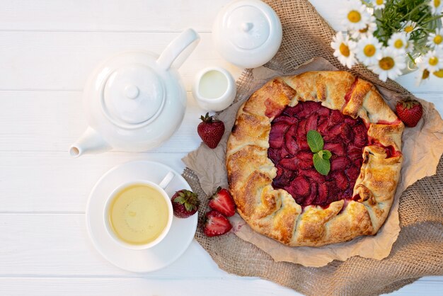 Recién horneado tarta casera de fresa galette, una taza de té, una tetera y un jarrón con un ramo de flores de margarita.