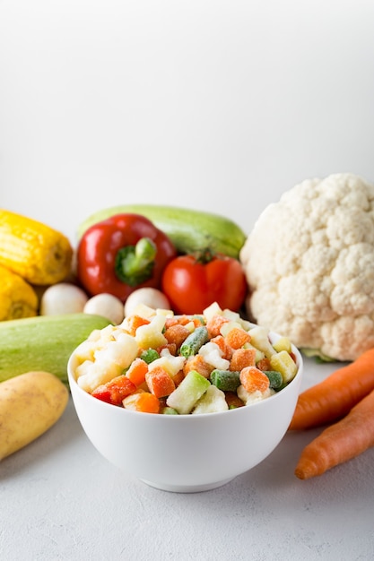 Recipiente de cerámica blanca con mezcla de verduras congeladas con lugar para texto sobre fondo gris y alimentos frescos