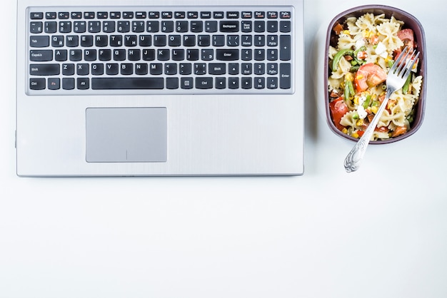 Recipiente con ensalada con pasta en el lugar de trabajo cerca de la computadora.