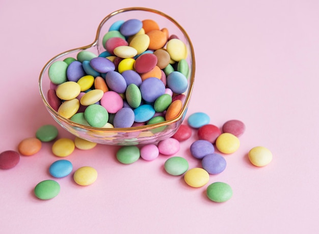 Recipiente en forma de corazón con dulces de grageas multicolores sobre una superficie rosa