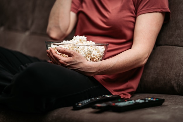 Foto recipiente con palomitas de maíz mientras ve una película con los controles remotos de la televisión en el sofá