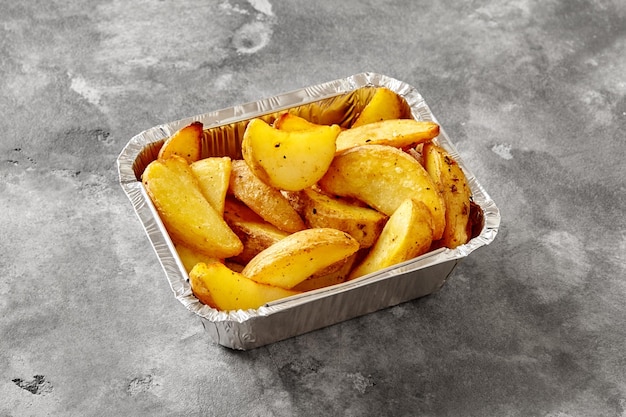 Recipiente de papel de aluminio con gajos de patata al horno sobre una mesa de piedra gris